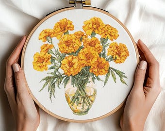 May include: Cross-stitch embroidery of a bouquet of yellow marigold flowers in a glass vase, set in a wooden hoop. The flowers are detailed with shades of yellow and orange, with green leaves. The background is white fabric.