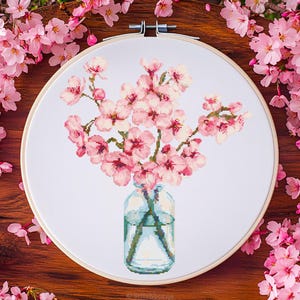 Könnte beinhalten: Kreuzstich-Stickerei mit rosa Kirschblüten in einer Glasvase, eingefasst in einem Holzreifen. Die Blüten sind rosa und weiß, mit einer transparenten Glasvase mit blauem Wasser. Der Hintergrund ist weiß.