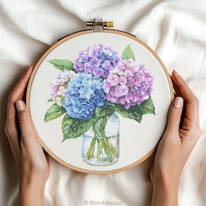 Puede incluir: Bordado de punto de cruz de un ramo de hortensias en un jarrón de cristal, sujeto en un bastidor de bordado de madera. Las flores son en tonos azules, morados y rosas con hojas verdes. El texto "Stitchborne" está en la parte inferior.