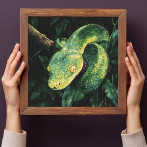 May include: A framed cross-stitch depicting a green snake coiled on a branch, set against dark green foliage. The snake features yellow eyes and a textured design. The wooden frame is held by hands.