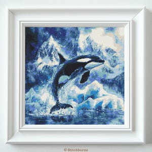 Peut inclure: Une œuvre d'art au point de croix encadrée représente une orque noire et blanche sautant hors de l'eau. L'arrière-plan présente des montagnes et des nuages bleus et blancs, créant une scène marine. L'œuvre est encadrée dans un cadre carré blanc.