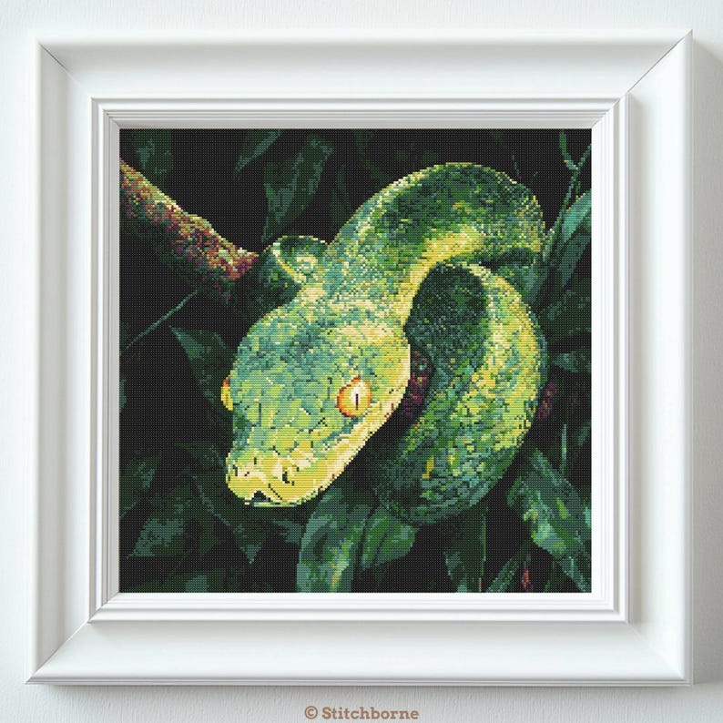 May include: A framed cross-stitch of a green snake coiled around a branch, surrounded by dark green leaves. The snake has yellow eyes and a yellow and green patterned body. The artwork is in a white frame.