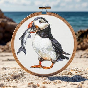 Op de afbeelding: Kruissteekborduurwerk van een papegaaiduiker die een vis in zijn bek houdt, geplaatst in een houten borduurring. De vogel heeft een zwart, wit en oranje verenkleed. De achtergrond toont een strand en de zee.
