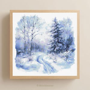 Pode incluir: Uma paisagem de inverno nevada em ponto cruz, emoldurada. A obra apresenta um caminho através de uma floresta coberta de neve com árvores nuas e sempre-verdes. A paleta de cores é principalmente azul e branco, com uma moldura de madeira castanha clara.