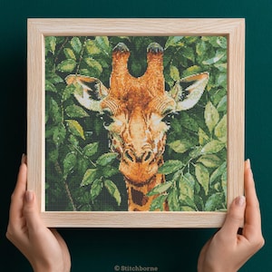 May include: A framed cross-stitch of a giraffe's face, set against a backdrop of green leaves. The giraffe has a brown and tan patterned coat, with large eyes and small ears. The frame is made of light-colored wood.