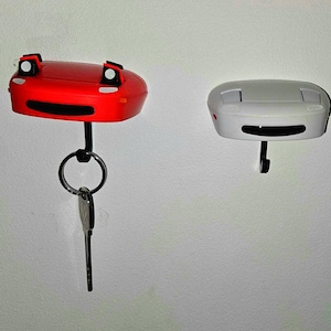 Puede incluir: Tres llaveros con forma de coche en rojo, blanco y rosa, cada uno con un gancho negro y un diseño de cara sonriente. El soporte rojo tiene una sola llave, mientras que el rosa contiene un juego de llaves. El blanco está vacío.