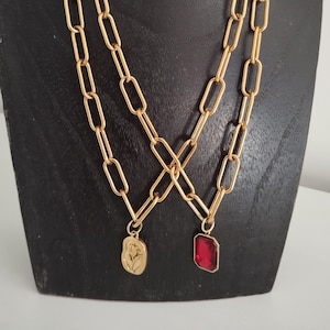 Pode incluir: Duas correntes douradas com elos retangulares. Uma corrente tem um pingente dourado com um desenho floral, e a outra tem um pingente de pedra preciosa vermelha retangular. As correntes são exibidas em um manequim preto.