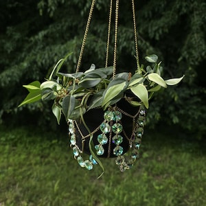 May include: A black hanging planter with a trailing green plant. The planter is suspended by gold chains and adorned with strands of iridescent, light green and blue crystal beads. The background is blurred green foliage and grass.