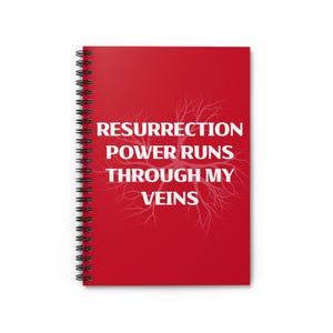 以下が含まれることがあります： 赤いスパイラル綴じノート。白い文字で「RESURRECTION POWER RUNS THROUGH MY VEINS」と書かれています。テキストは垂直に配置され、背景には木の枝のデザインがあります。