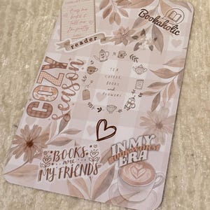 Op de afbeelding: Een decoratieve kaart met een gezellig thema, met tekst en illustraties in bruin- en beigetinten. De kaart bevat zinnen als "Cozy Season", "Books are my friends" en "In my bookworm era". Het toont ook theekopjes, bloemen en een kopje koffie.