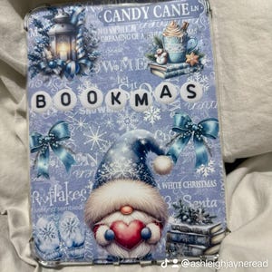 Könnte beinhalten: Eine hellblaue Tablet-Hülle mit Winterthema. Die Hülle zeigt einen Zwerg, der ein Herz hält, Schneeflocken, Schleifen und das Wort "BOOKMAS". Weitere Texte sind "Candy Cane Ln" und "A White Christmas".