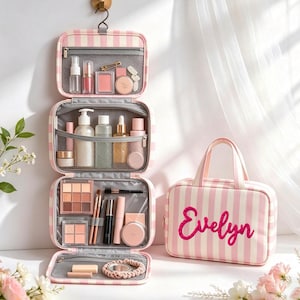 Puede incluir: Un neceser colgante con rayas verticales rosas y blancas, que presenta el nombre Evelyn bordado en letra cursiva rosa oscuro. El neceser está abierto para mostrar múltiples compartimentos de malla transparente llenos de productos de belleza.