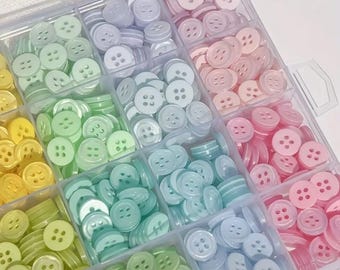 May include: A plastic storage box filled with various colors of small, round buttons. The buttons are arranged in rows and columns, and they are all the same size. The colors of the buttons include light blue, light green, light yellow, and light pink.