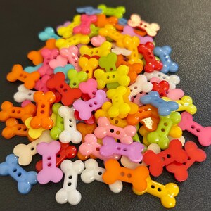 Könnte beinhalten: Ein Haufen bunter, knochenförmiger Knöpfe in verschiedenen Farbtönen, darunter Orange, Gelb, Rosa, Grün, Rot und Blau. Diese Kunststoffknöpfe haben zwei Löcher zum Annähen und eignen sich ideal für Bastelprojekte.
