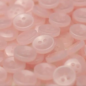 Pode incluir: Uma coleção de botões redondos rosa pálido com dois ou quatro furos. Os botões estão espalhados, criando uma superfície texturizada. Os botões são feitos de um material translúcido.