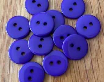 May include: A collection of glossy, round, purple buttons with two holes each. The buttons are scattered on a light brown wooden surface. The buttons are a solid, vibrant purple color.