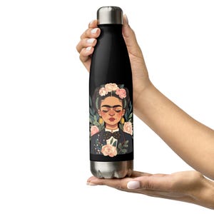 May include: Black insulated water bottle featuring a portrait of Frida Kahlo with a floral crown and a dark suit. The design includes pink and yellow flowers and green foliage. The bottle has a silver cap and base.