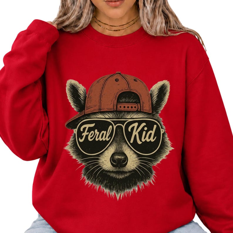 May include: A bright red crewneck sweatshirt featuring a graphic illustration of a raccoon wearing a brown baseball cap and aviator sunglasses. The text Feral Kid is printed in a stylised font across the lenses of the sunglasses on the raccoon.