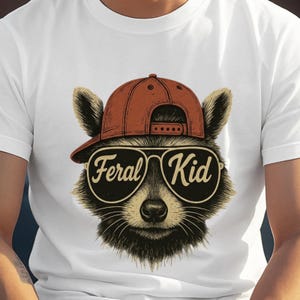 May include: A white t-shirt featuring a graphic illustration of a raccoon wearing a rust-coloured baseball cap and large aviator-style sunglasses. The lenses of the sunglasses display the text Feral Kid in a cursive font across the centre.