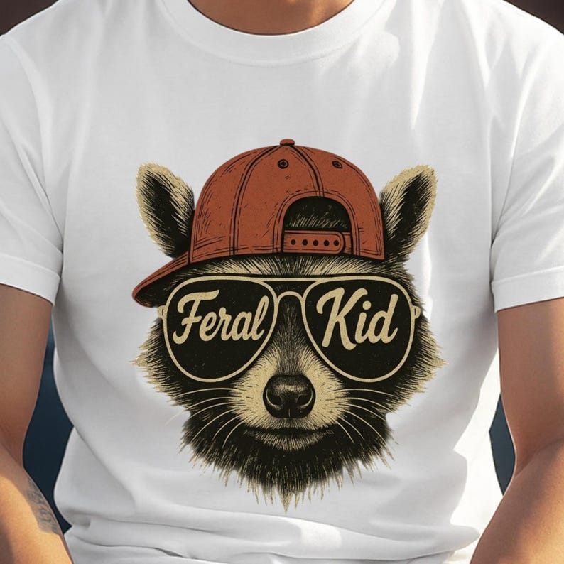May include: A white t-shirt featuring a graphic illustration of a raccoon wearing a rust-coloured baseball cap and large aviator-style sunglasses. The lenses of the sunglasses display the text Feral Kid in a cursive font across the centre.