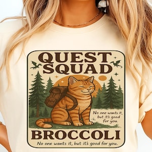 Könnte beinhalten: Cremefarbenes T-Shirt mit einer Grafik einer lächelnden orangefarbenen Tabby-Katze mit Rucksack, vor einer Waldszene. Der Text lautet "QUEST SQUAD" oben und "BROCCOLI" unten, mit dem Satz "No one wants it, but it's good for you."