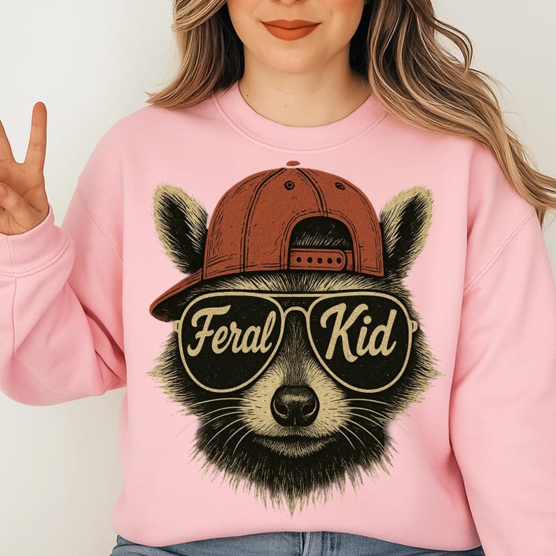 May include: A light pink crewneck sweatshirt featuring a graphic illustration of a raccoon wearing a brown baseball cap and large aviator sunglasses. The text "Feral Kid" is printed in a stylised font across the lenses of the sunglasses.