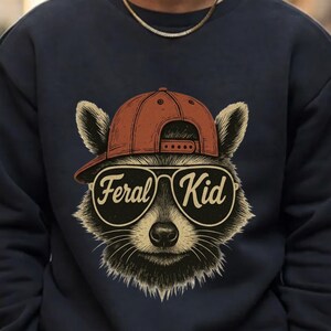 May include: A navy blue sweatshirt featuring a graphic illustration of a raccoon wearing a rust-coloured baseball cap and large aviator-style sunglasses. The lenses of the sunglasses display the white cursive text Feral Kid across the centre.