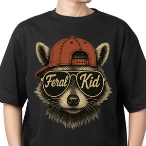 May include: A black t-shirt featuring a graphic illustration of a raccoon wearing a red baseball cap and aviator sunglasses. The lenses of the sunglasses display the text Feral Kid in a stylised script font. The raccoon has detailed fur.
