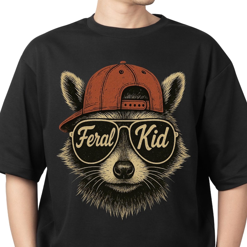 May include: A black t-shirt featuring a graphic illustration of a raccoon wearing a red baseball cap and aviator sunglasses. The lenses of the sunglasses display the text Feral Kid in a stylised script font. The raccoon has detailed fur.
