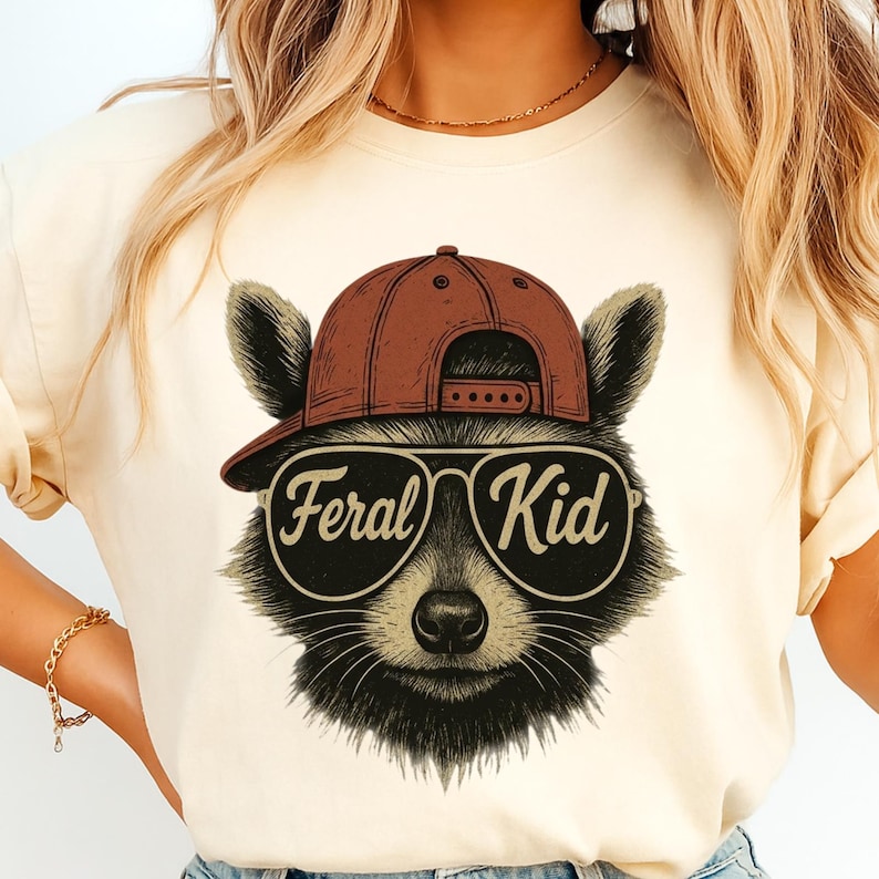 May include: A cream-coloured t-shirt featuring a graphic of a raccoon wearing a brown baseball cap and aviator sunglasses. The lenses of the sunglasses display the text Feral Kid in a cursive font. The raccoon has black and grey fur.