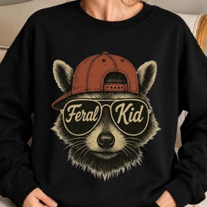 May include: A black crewneck sweatshirt featuring a graphic of a raccoon wearing a rust-coloured baseball cap and aviator sunglasses. The lenses of the sunglasses display the text Feral Kid in a cursive font, centred on the chest of the garment.