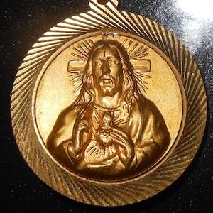 May include: A gold-colored pendant featuring a detailed relief of Jesus Christ. The pendant has a circular design with a textured border and a loop for hanging. The image of Jesus includes a cross and a heart.
