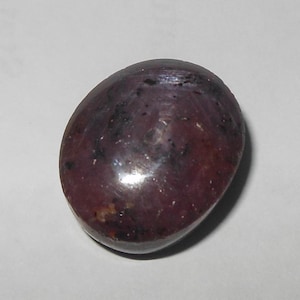 May include: An oval-shaped cabochon gemstone with a deep, dark reddish-purple hue. The surface of the stone displays a smooth, polished finish with visible natural inclusions and dark speckles throughout its opaque, rounded body structure.