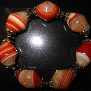 May include: A beaded bracelet featuring large, polished agate stones in shades of red, white, and tan. The stones are separated by ornate metal beads and dark brown spacers. The bracelet is a striking piece of jewelry.