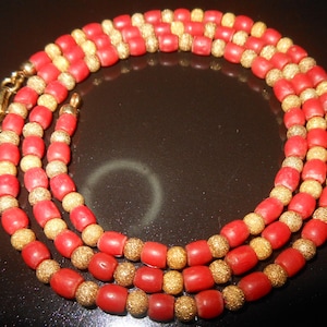May include: A three-strand necklace featuring alternating red and gold-colored beads. The red beads are barrel-shaped, while the gold beads have a textured, glittery appearance. The necklace is secured with a gold-colored clasp.