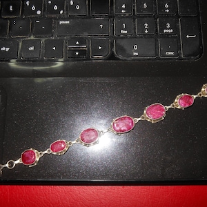 Könnte beinhalten: Ein silbernes Armband mit ovalen, tiefroten Edelsteinen. Das Armband besteht aus einzelnen Gliedern, die die Steine verbinden, mit einem Verschluss. Der Schmuck wird auf einer schwarzen Laptop-Oberfläche präsentiert.