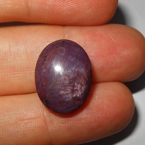 May include: An oval-shaped, cabochon-cut gemstone with a deep purple and reddish-maroon hue. The stone features a smooth, polished surface with visible internal inclusions and natural color variations throughout its translucent body.