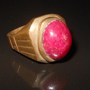 May include: A gold-colored ring with a large, oval, red gemstone. The ring has a textured band with etched lines and a raised bezel setting around the stone. The gemstone is a vibrant, deep red color.