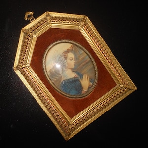 May include: An antique framed portrait featuring a woman in profile with clasped hands. The oval portrait is set against a red velvet background within a gold filigree frame. The frame has an ornate design and a decorative top loop.