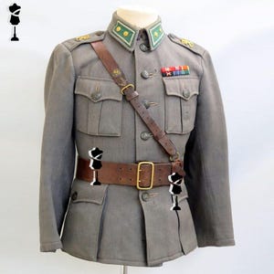 Könnte beinhalten: Eine graue Militärjacke mit Stehkragen, grünen und goldenen Abzeichen, vier Vordertaschen mit Knopfverschluss, einem braunen Ledergürtel mit Messingschnalle und einem passenden braunen Leder-Schulterriemen über der Brust.