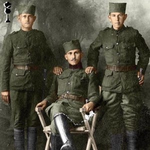 May include: A vintage photograph of three men in olive green military uniforms and caps. The man in the centre is seated in a wooden chair, while the other two stand on either side. All three wear brown belts and boots.