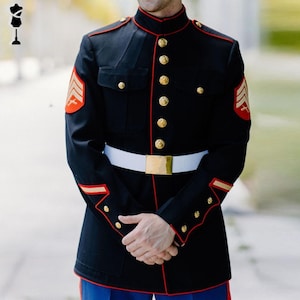 May include: A dark navy blue military uniform with gold buttons, red trim, and a white belt with a gold buckle. The uniform features red and gold insignia on the sleeves. The person is wearing blue trousers.