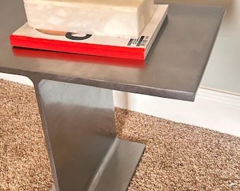 Op de afbeelding: Een zilveren metalen tafel met een rechthoekig blad en een enkel I-balkpoot. Een witte doos met een schedeldeksel staat op een rood boek op de tafel.