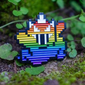 May include: A pixelated enamel pin featuring a colorful design with a rainbow gradient. The pin has a white and red top with black accents and is set against a backdrop of green moss and foliage.