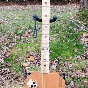 May include: A handmade cigar box guitar with a wooden neck and a brown cigar box body. The box features a skull and crossbones design, along with the brand name "Padron". The guitar is supported by a black stand.