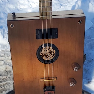 May include: A handmade wooden cigar box guitar with a light brown finish. The guitar features a Celtic knot design on the sound hole and a cross design on the bridge. It has four strings and tuning pegs.