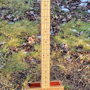 May include: A handmade cigar box guitar with a wooden neck and a red and gold box. The guitar has strings and tuning pegs. The box reads "30th Anniversary". The guitar is resting on a metal stand.