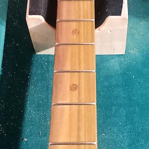 May include: A guitar neck with a light wood finish, featuring inlaid fret markers. The neck is supported by a wooden stand with black padding. The background is a teal-colored surface.