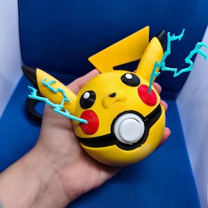 May include: A yellow Pikachu-shaped 3D-printed object with black accents, red cheeks, and blue lightning bolt details. The object has a white button and a black band around the middle. The ears are black-tipped.