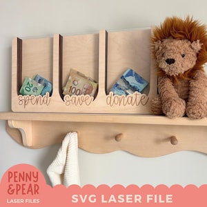 Puede incluir: Organizador de dinero de madera con tres compartimentos etiquetados como "gastar", "ahorrar" y "donar". Cada sección contiene billetes. Un peluche de león está a la derecha. La estantería tiene un borde decorativo y clavijas.